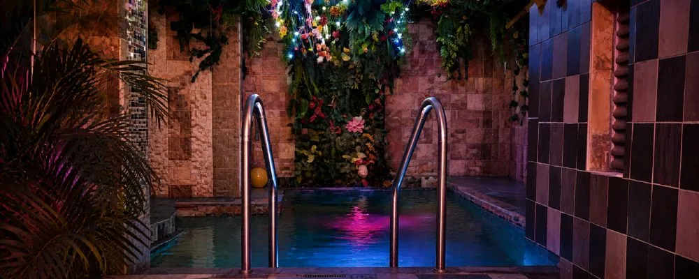 Zonas húmedas de Club SW69: piscina iluminada en azul y rosa, con muro de piedra, jardín vertical y barandas de acero