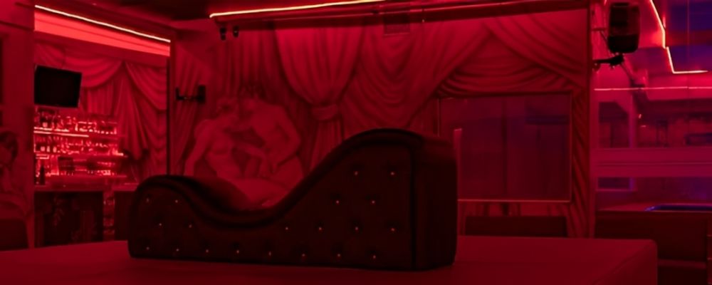 Sala de Fantasía de Imperio SW: diván tántrico oscuro frente a un mural greco-romano, bajo luz roja