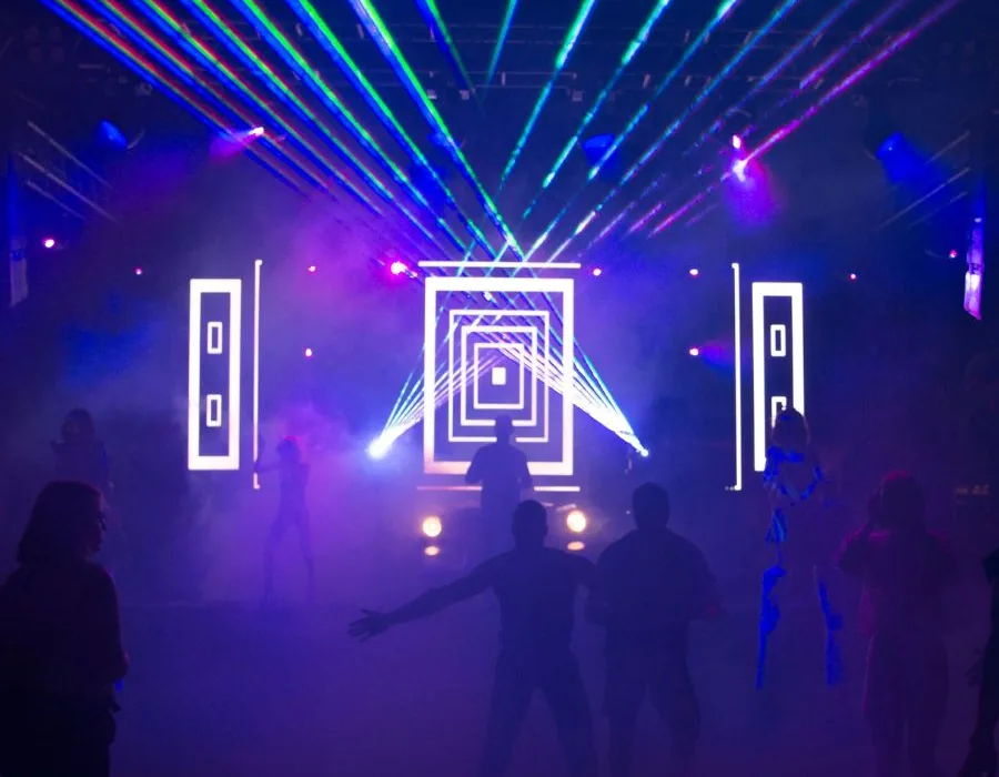 Pista de baile de un club nocturno con haces de láser verdes y azules sobre el humo y siluetas de personas bailando