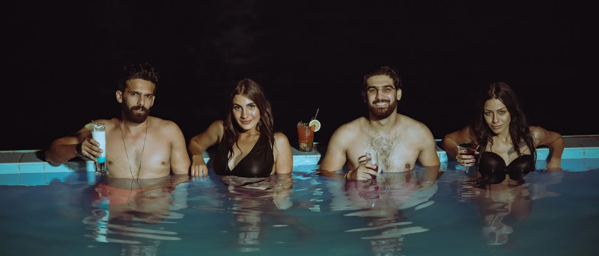 Dos parejas swinger compartiendo cócteles en una piscina de noche