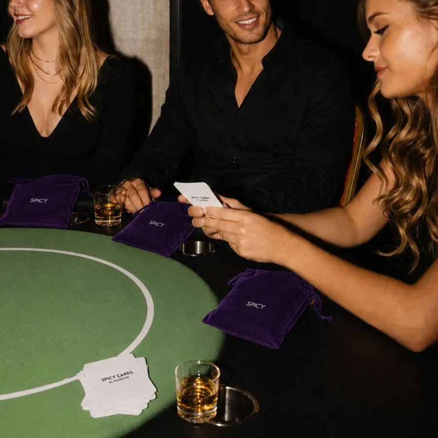 Tres personas jugando Spicy Cards alrededor de una mesa de paño verde, con bolsas de terciopelo morado y vasos servidos